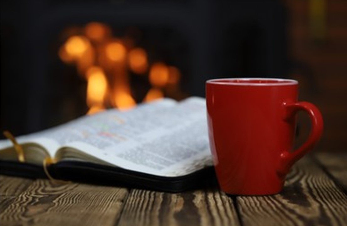 St. John's United Church | A red mug sits on a wooden table next to an open book. A fireplace with burning flames is blurred in the background.