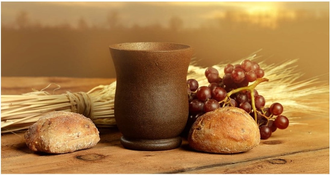 St. John's United Church | A rustic table displays a wooden cup, red grapes, two bread rolls, and a bundle of wheat, with a sunset in the background.