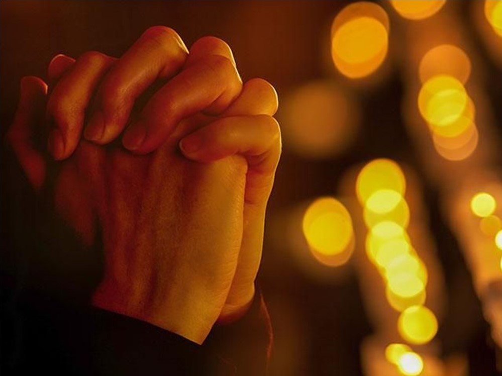 St. John's United Church | Hands clasped together in front of blurred warm golden lights.