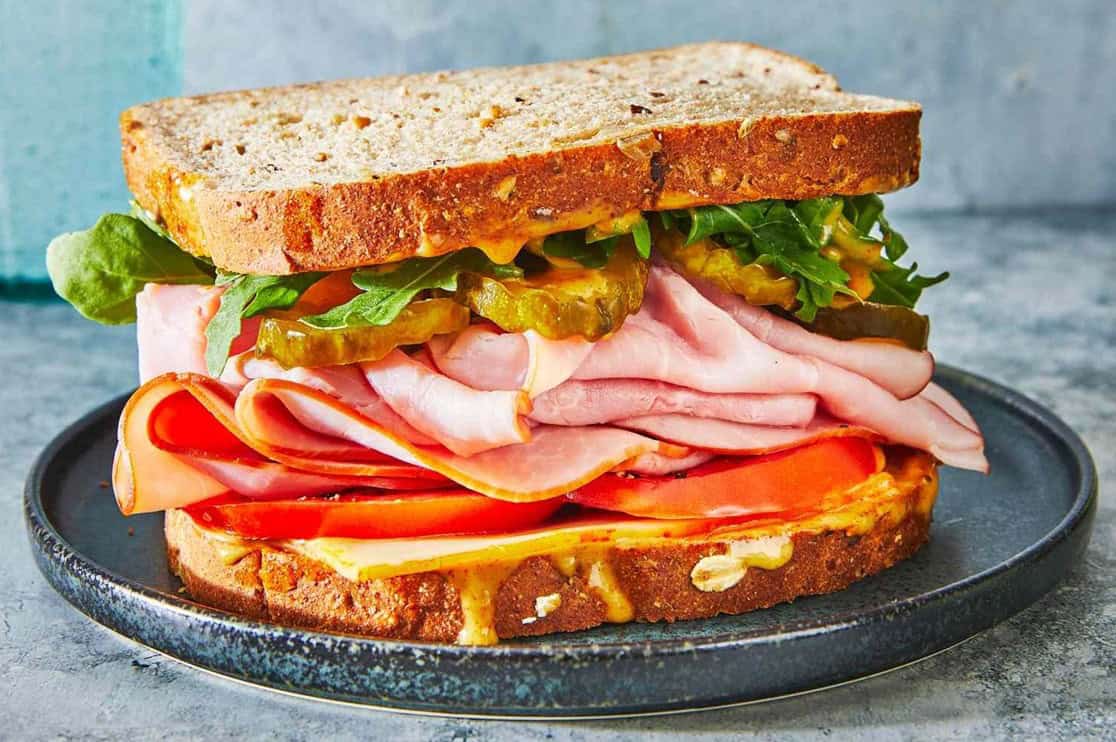 St. John's United Church | A ham sandwich on whole grain bread with lettuce, pickles, tomato, and mustard, served on a dark plate.