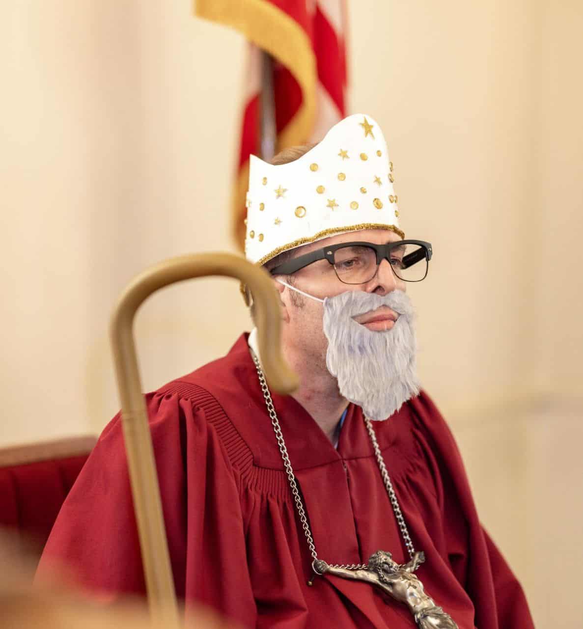St. John's United Church | A person dressed in a maroon robe, wearing a white and gold hat, gray fake beard, glasses, and a large cross necklace sits in front of a U.S. flag, reflecting the beliefs and teachings at St. John's United Church.