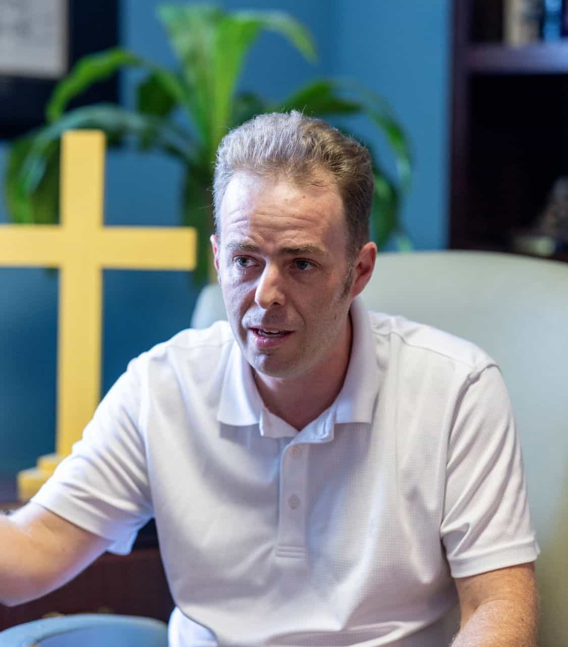 St. John's United Church | A man in a white polo shirt sits and gestures while talking, with a large yellow cross and a green plant in the background, sharing insights into the beliefs and teachings at St. John's United Church.